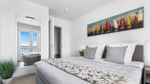 Ein modernes Schlafzimmer mit einem großen Bett, einem Segelbootgemälde, einer Topfpflanze und Blick durch eine offene Tür.