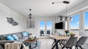 Ein modernes Wohnzimmer mit grauem Sofa, einem Couchtisch und einem Essbereich mit Holztisch und Stühlen. Ein großes Fenster bietet Blick auf das Meer, und an der Wand ist ein Fernseher angebracht.