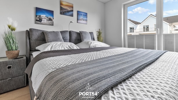 Ein modernes Schlafzimmer mit einem großen Bett, grau-weißer Bettwäsche, einer grauen Kopfteil und zwei an der Wand hängenden Bildern.