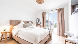 Ein modernes Schlafzimmer mit einem großen Bett, einem Nachttisch mit Lampe, einer kleinen Pflanze und Blick ins Freie durch ein großes Fenster.