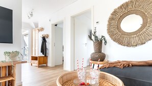 Ein modernes Wohnzimmer mit Holzboden, einem runden Gewebespiegel, einem blauen Sofa, einem Holztisch mit Gläsern und einer Obstschale sowie einer Tür zu einem anderen Raum.