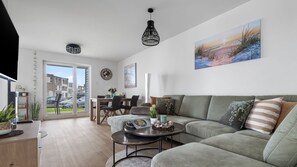 Un salon moderne avec un canapé, une table basse et un coin repas. Un tableau orne le mur et une vue sur l’extérieur est visible.