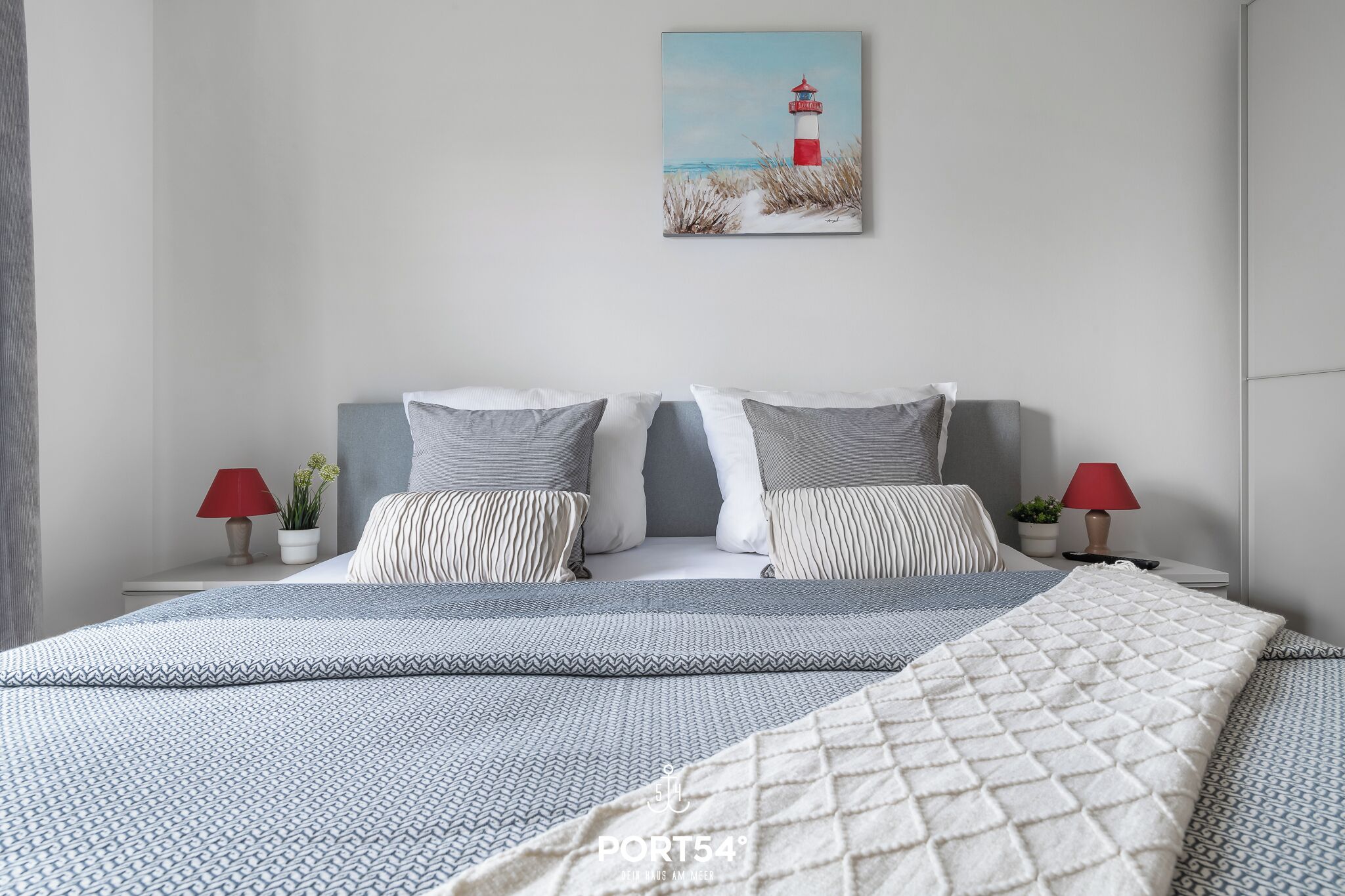Een netjes opgemaakt bed met een geborduurde sprei en kussens, een schilderij van een vuurtoren aan de muur, en twee nachtkastjes met lampen en kleine planten.