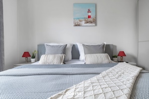 Un lit bien fait, recouvert d’une couvre-lit à motifs et agrémenté d’oreillers, un tableau représentant un phare accroché au mur, et deux tables de chevet ornées de lampes et de petites plantes.