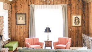 A cozy room with wooden walls, a green sofa, two pink armchairs, a small table with a lamp, and framed pictures on the walls.