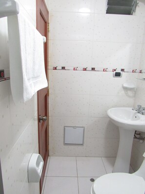 Un baño con un lavabo blanco, un inodoro, una toalla colgada en un perchero y una pequeña ventana sobre el lavabo.