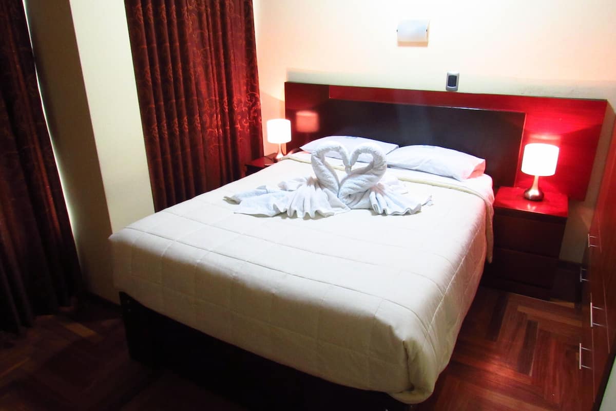 A hotel room with a neatly made bed, a red headboard, two bedside lamps, and a wooden floor.