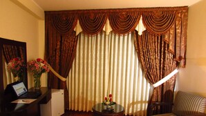Una habitación con piso de madera, una mesita con un florero, una silla, un escritorio con una computadora portátil y un ventanal con cortinas.