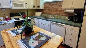 A kitchen with white cabinets, a brick wall, a wooden table with a placemat, a potted plant, and a microwave.