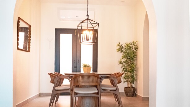 A dining area with a round table, six chairs, a hanging light fixture, and a potted plant.