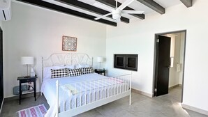 A bedroom with a white bed, a ceiling fan, a nightstand with a lamp, a decorative wall piece, and a door leading to another room.