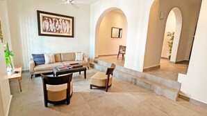 A spacious living area with a sofa, coffee table, and chairs, a ceiling fan, and a framed artwork on the wall.