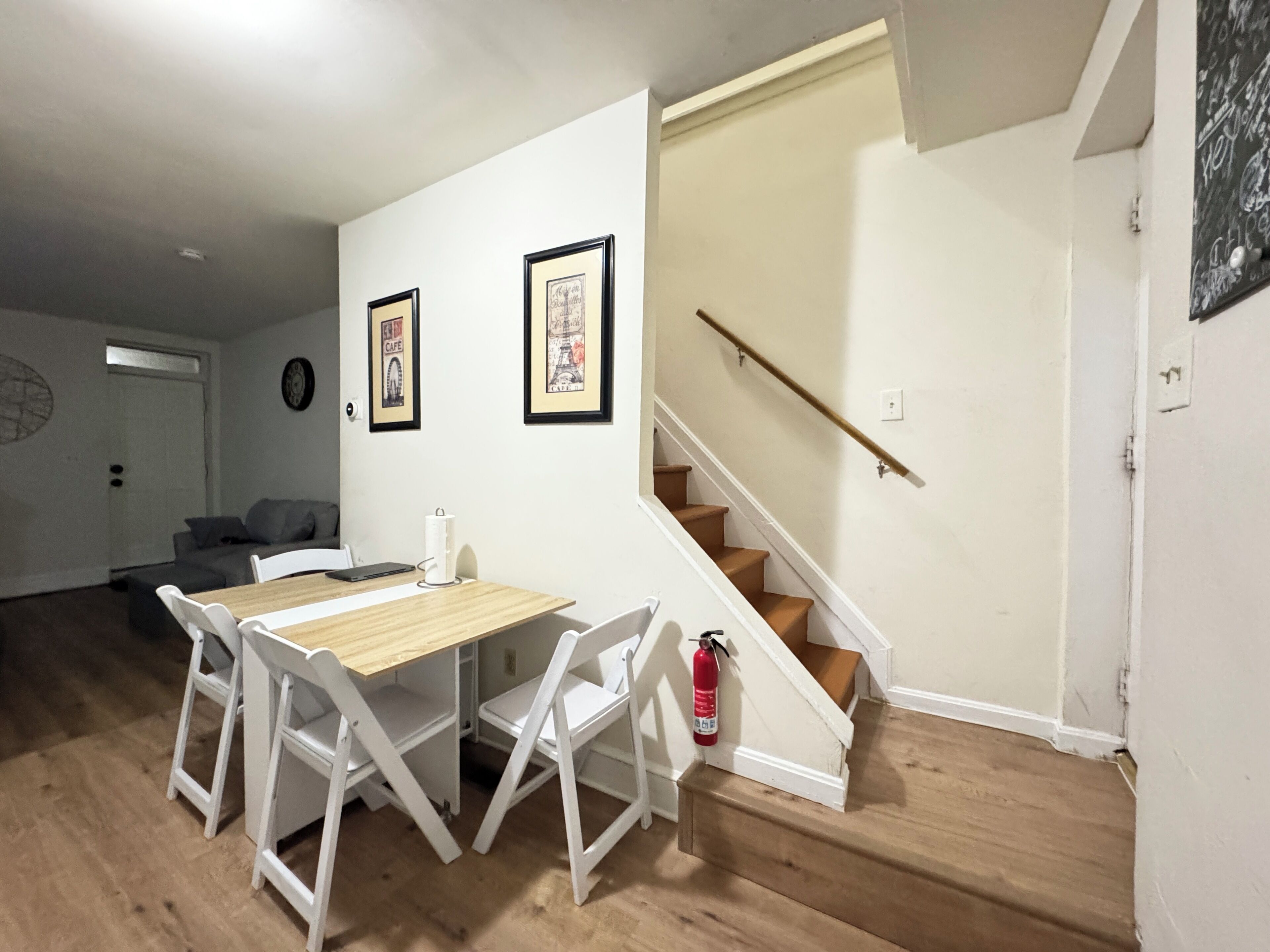 A dining area with a wooden table and white chairs, a staircase with a handrail, and two framed pictures on the wall.