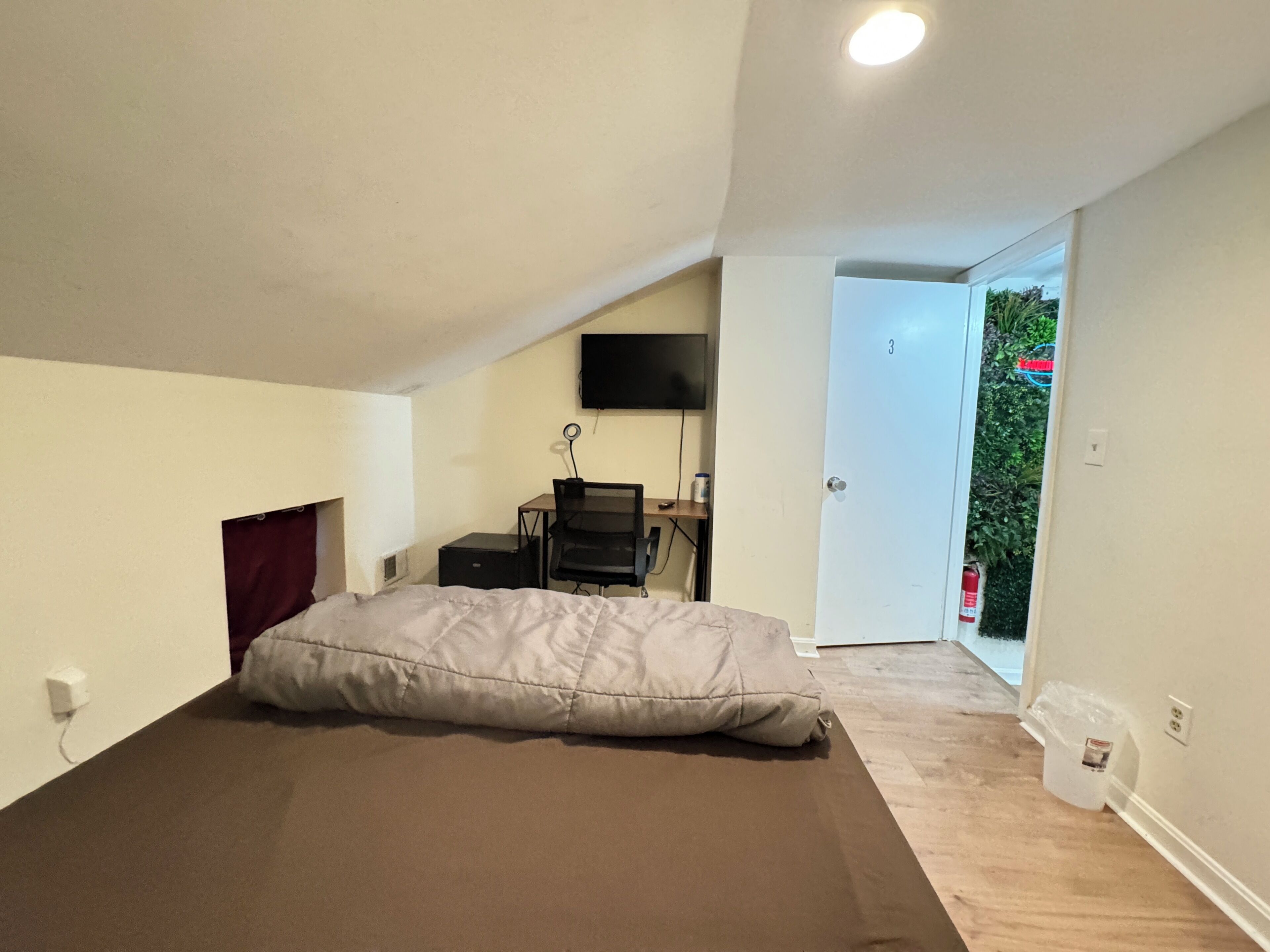 A bedroom with a bed, a desk with a chair, a TV on the wall, and a door leading to a balcony.