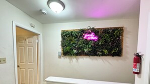 Un pasillo con una puerta blanca, un letrero de neón, un extintor y un conjunto de plantas en la pared.