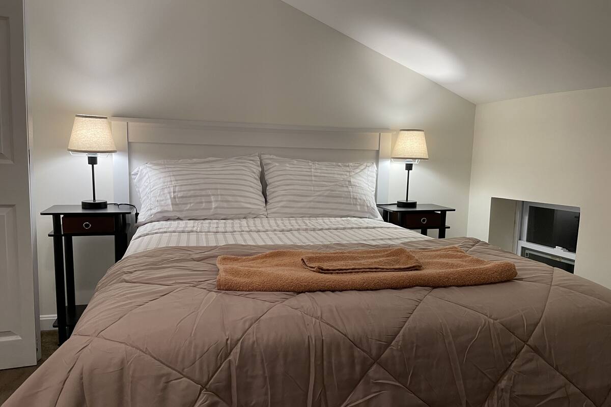 A neatly made bed with a brown quilt, two bedside lamps, and a television on a stand to the right.