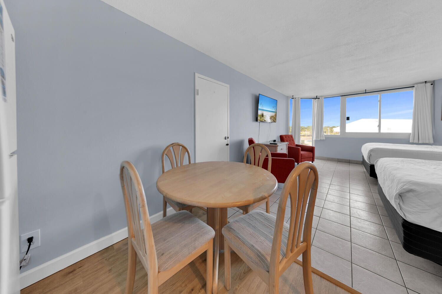 A room with a round wooden table, four chairs, a bed, a TV, and a window with curtains.