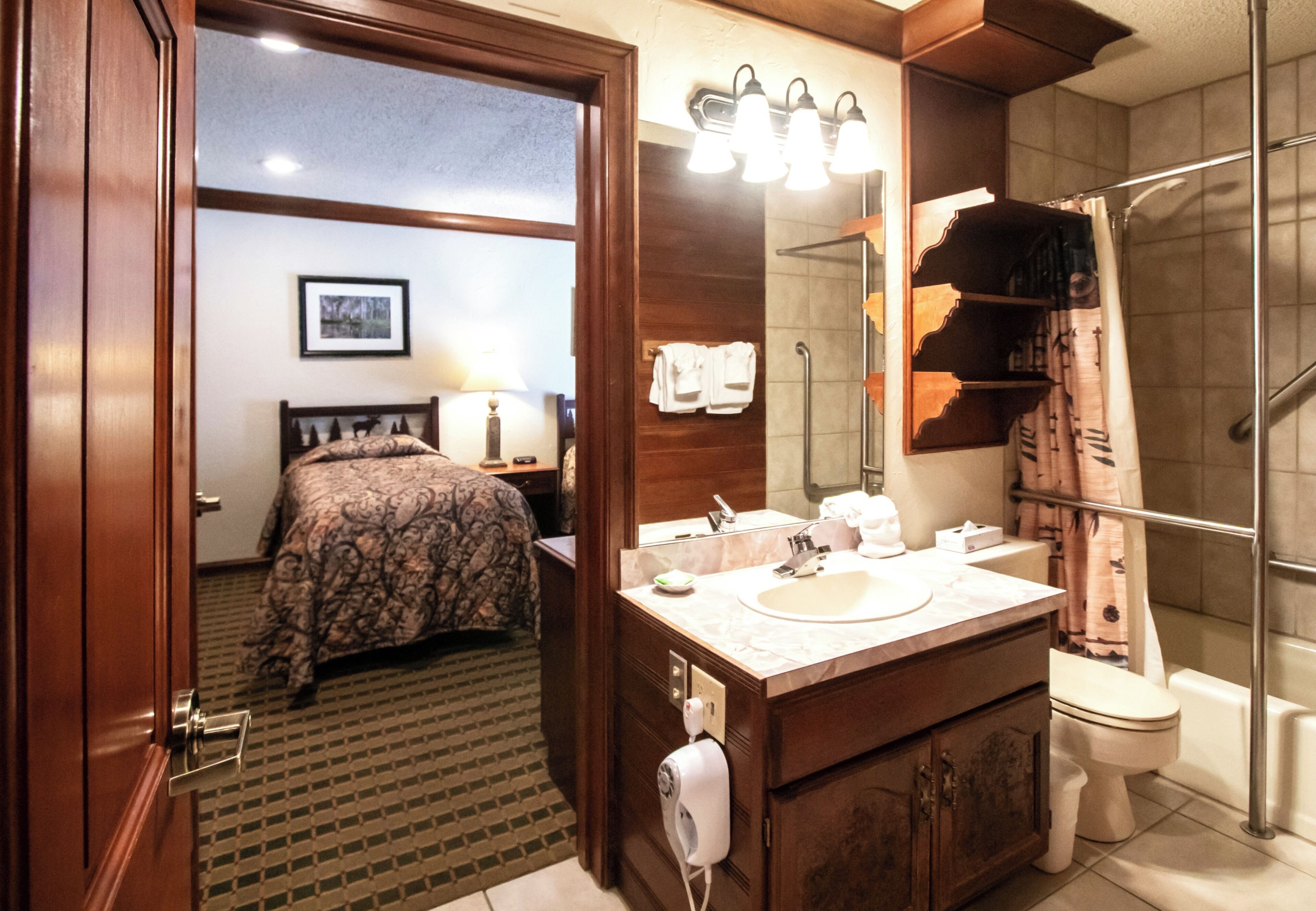 A hotel room with a wooden door, a bed with patterned bedding, a bathroom with a sink, mirror, and shower.