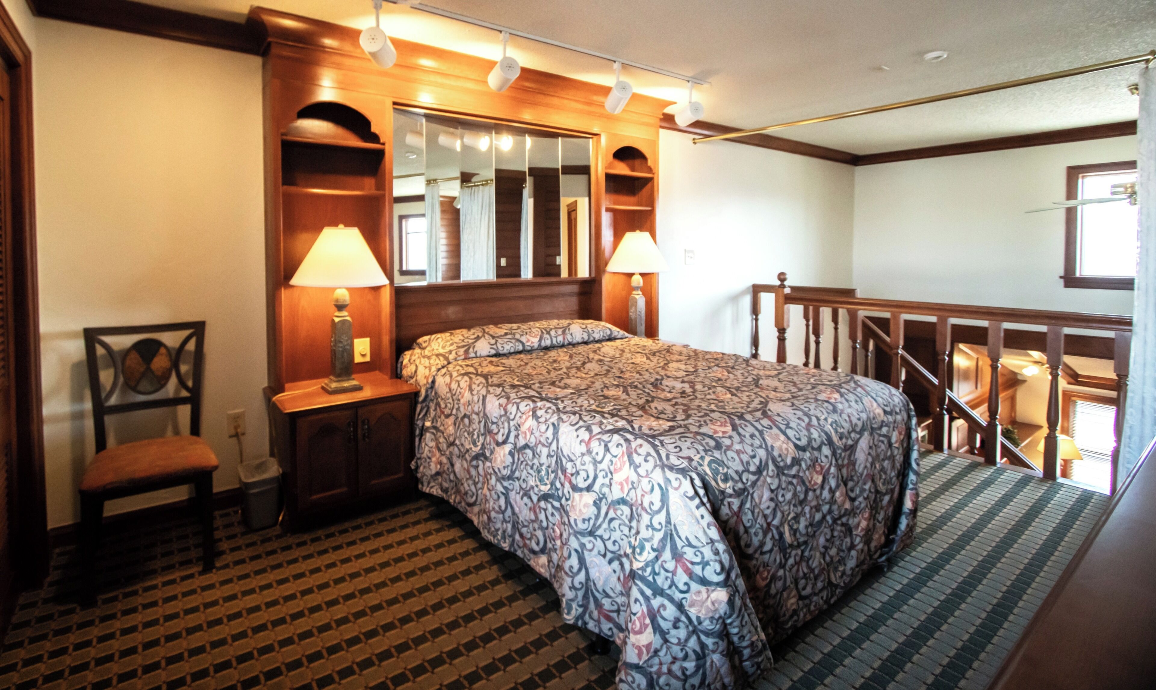 A hotel room with a large bed, two bedside lamps, a wooden headboard, a small chair, a trash bin, a patterned rug, and a view of the balcony.