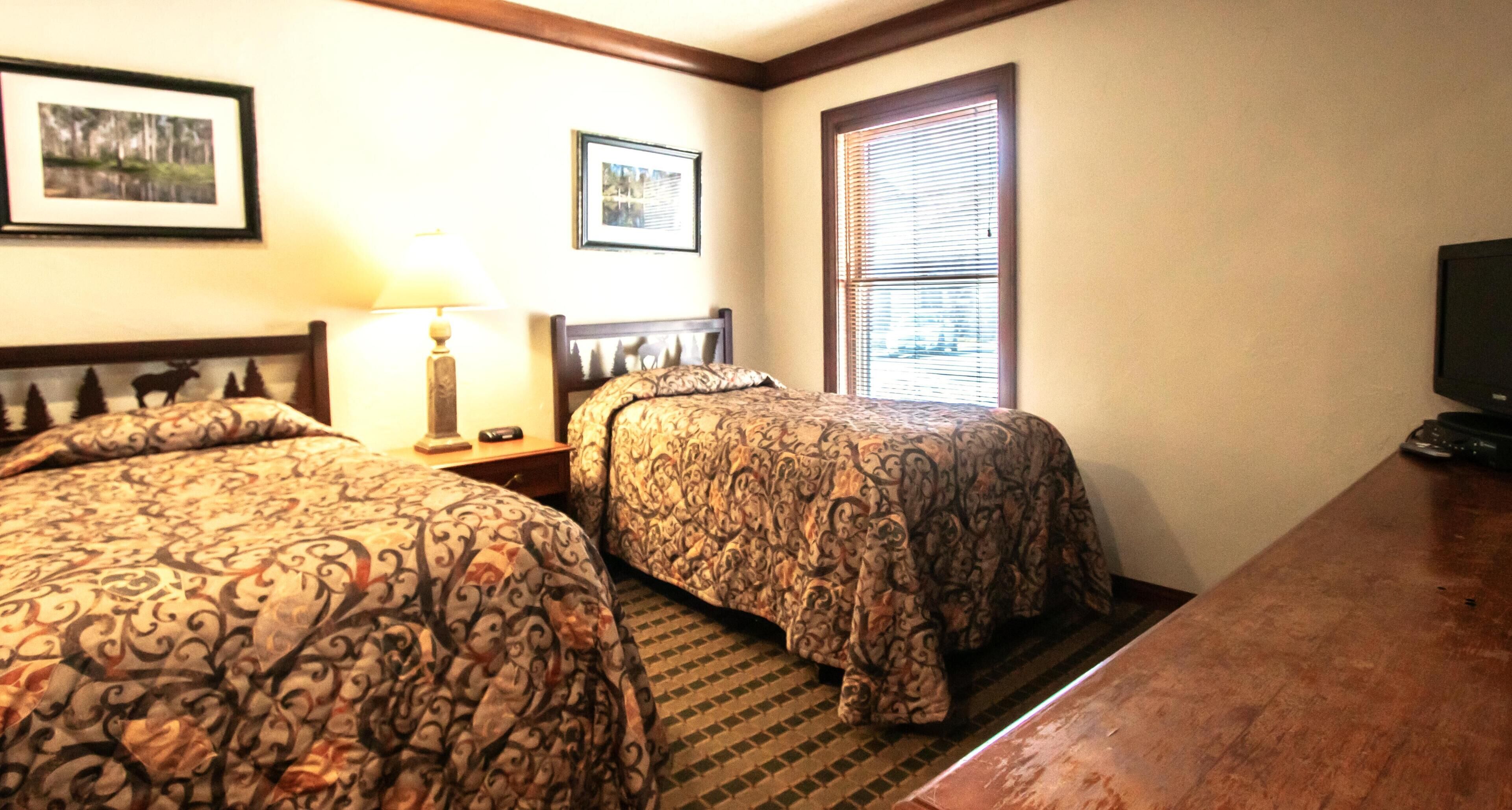 A hotel room with two beds, a nightstand, a lamp, a television, and a window with blinds.