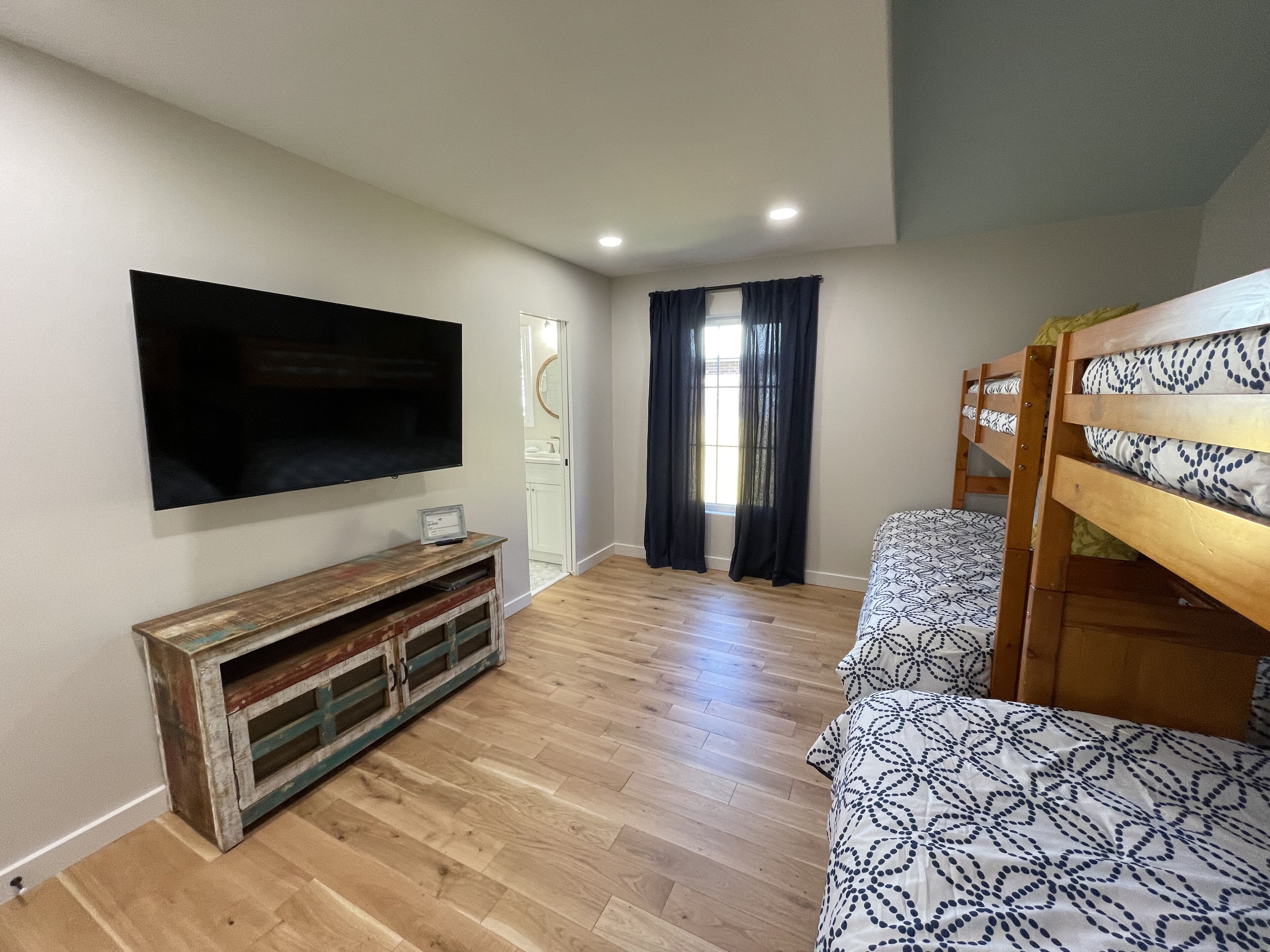 A room with a TV mounted on the wall, a wooden bench, bunk beds, and a window with curtains.