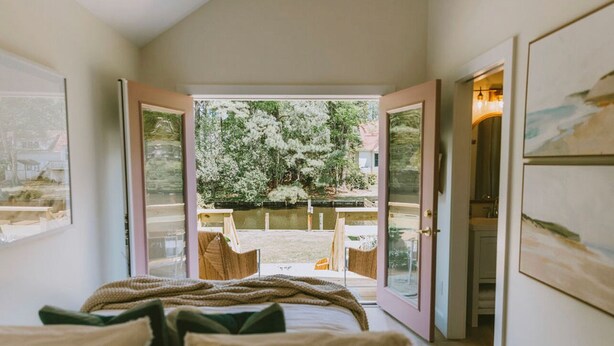 A bedroom with a bed, a view of a garden with trees and a pond, and a door leading to a bathroom. - Canal front, Modern Tiny House! Bikes. Fire pit! (Kitty Hawk)