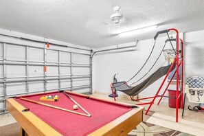 Un tavolo da biliardo con le sue bocce, un'amaca e una scala rossa in una stanza con soffitto e pareti bianche.