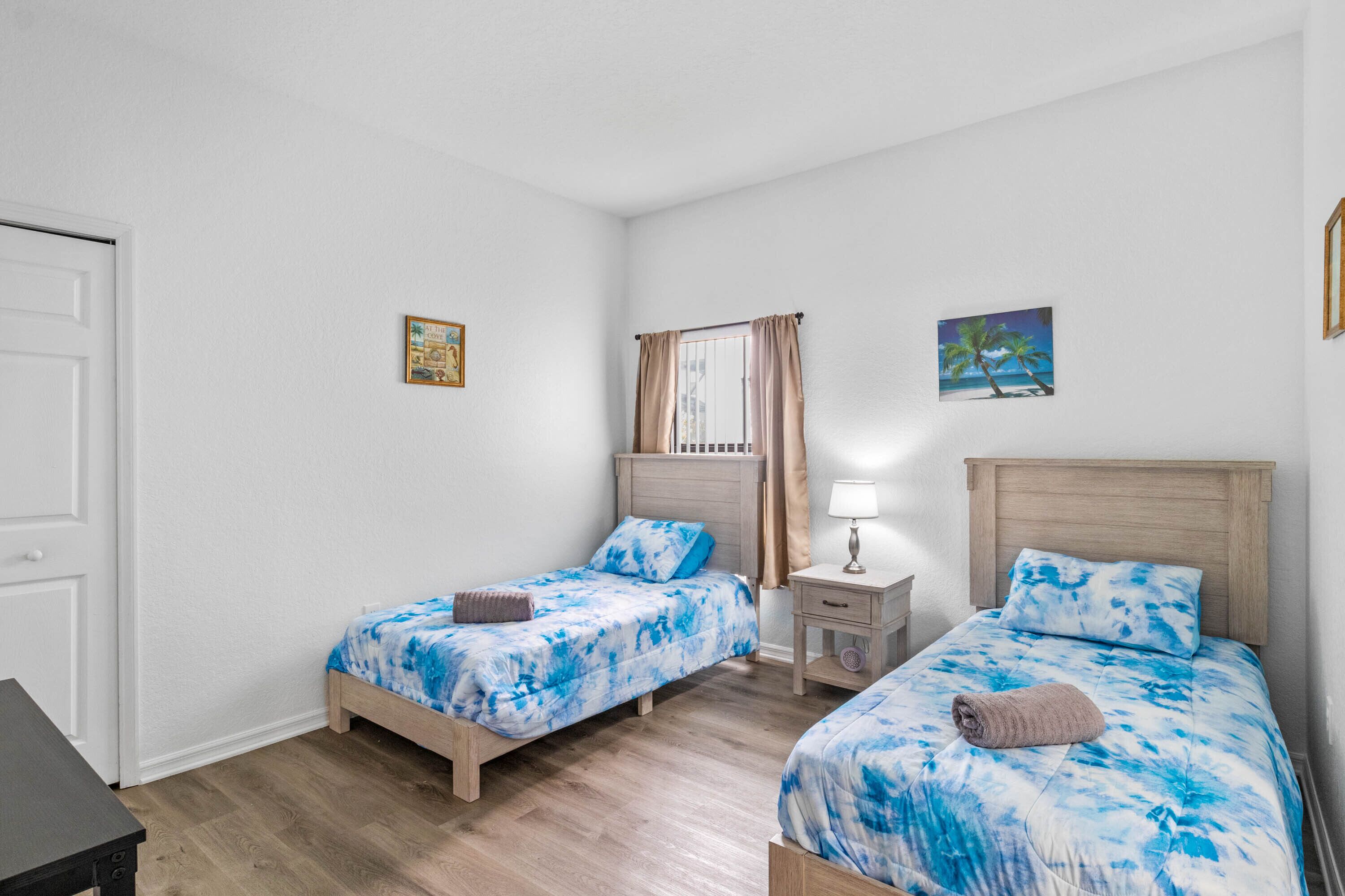 A room with two single beds, a nightstand with a lamp, a door, and a small table.