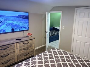 Una habitación con un televisor sobre un tocador de madera, un tapete estampado, una puerta que da a un dormitorio y una puerta blanca.