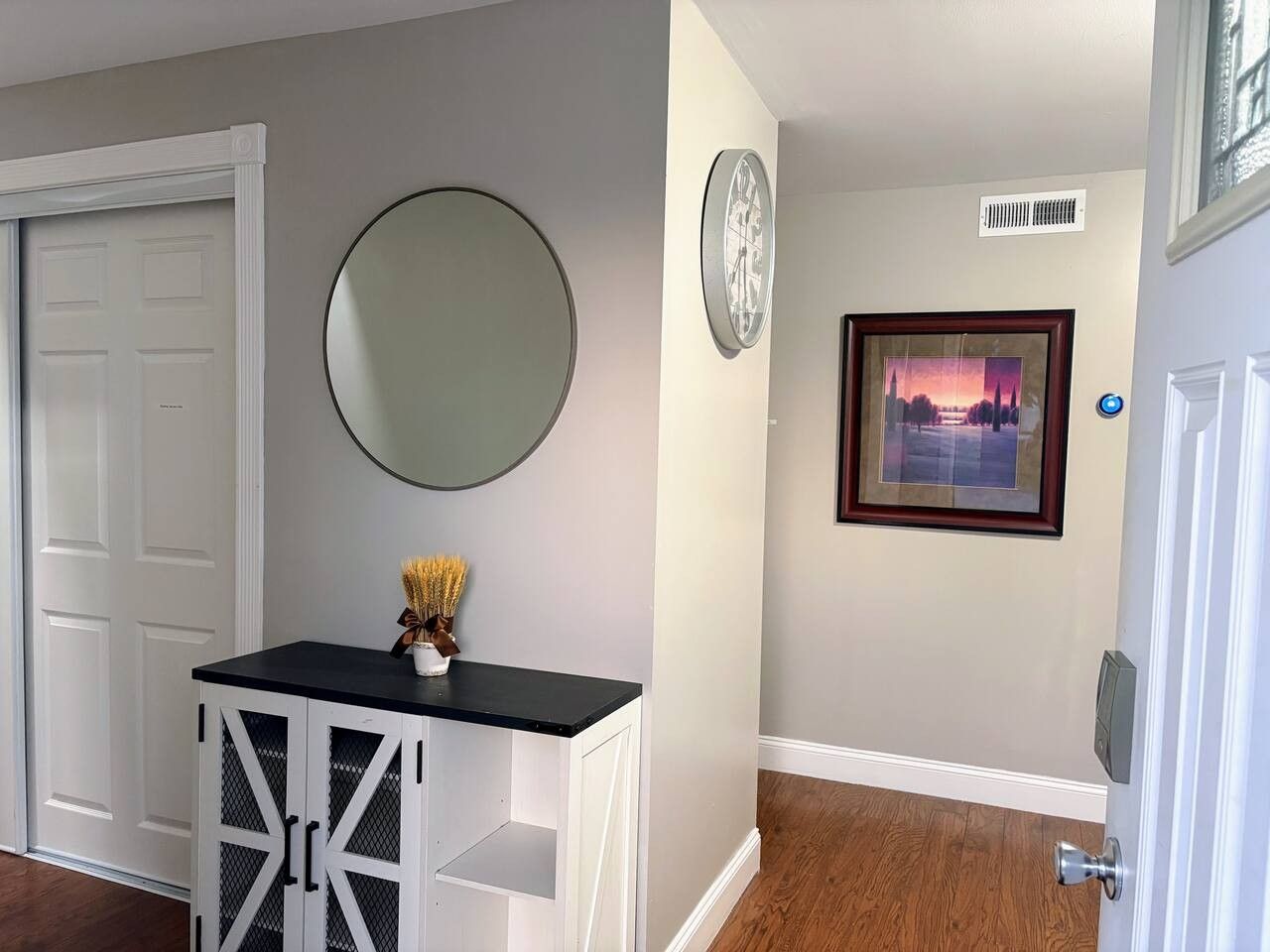 A hallway with a white door, a round mirror, a framed picture, and a small table with a vase.