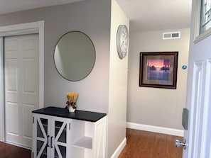 A hallway with a white door, a round mirror, a framed picture, and a small table with a vase. - Spacious 3BR Home | Recently Remodeled | Private Yard & Parking (West Seneca)