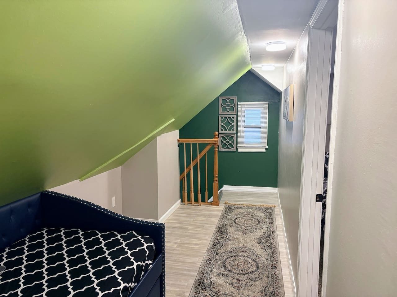 A hallway with a wooden staircase, a patterned rug, and a door leading to a room with green walls.