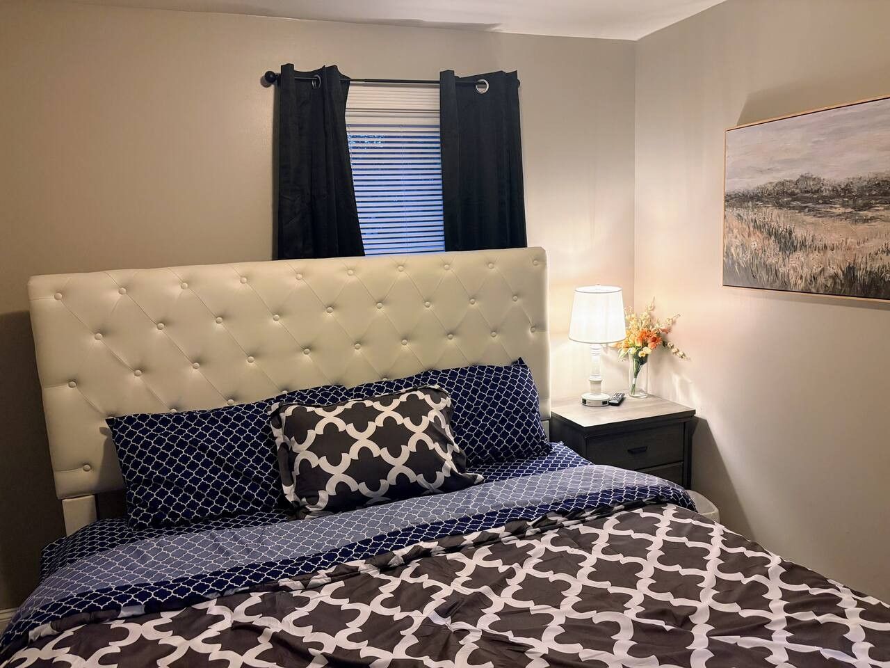 Une chambre à coucher avec une tête de lit capitonnée, une literie à motifs, une table de chevet avec une lampe et des fleurs, et un tableau accroché au mur.