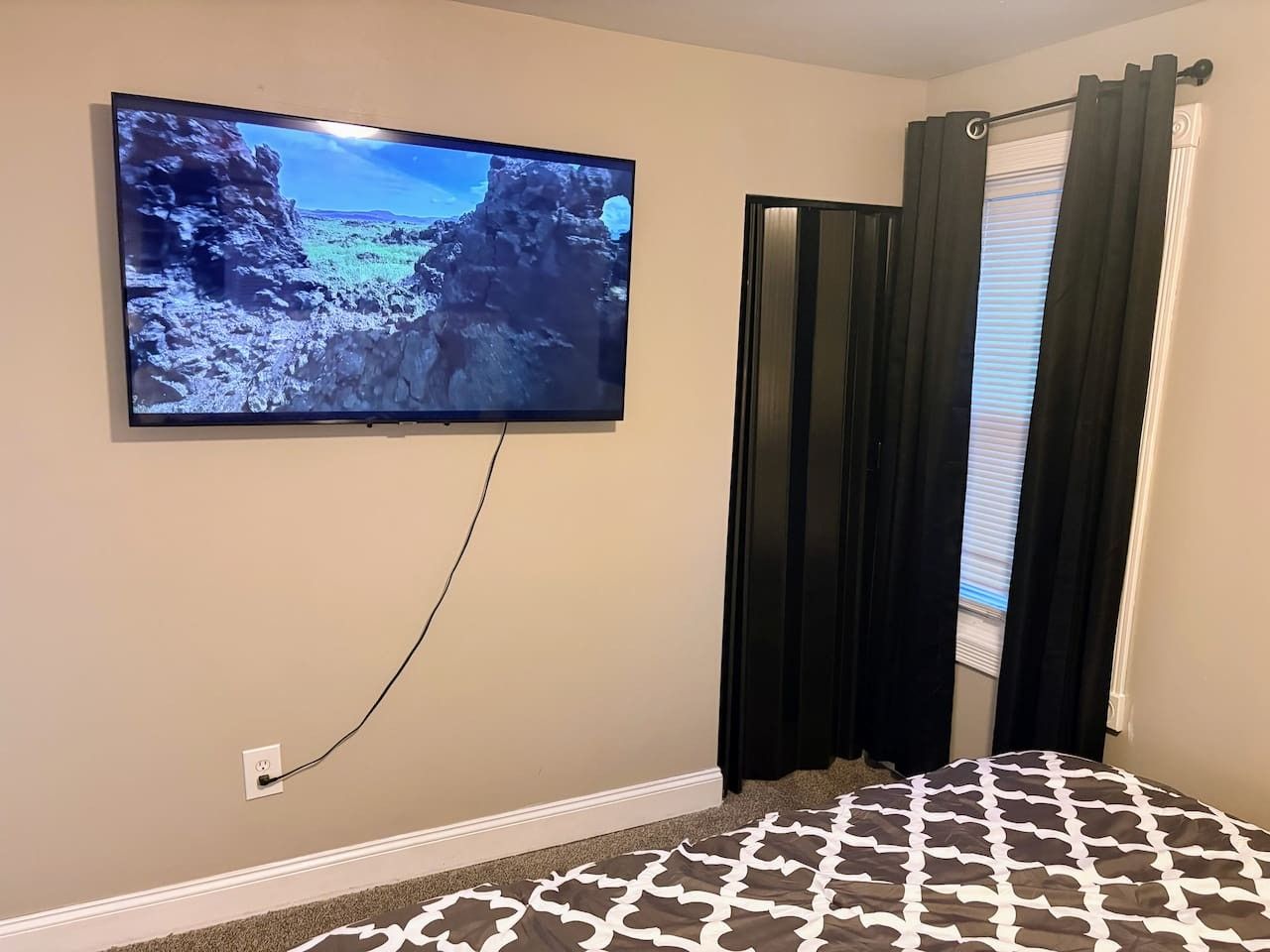Una habitación con un televisor montado en la pared, una ventana con cortinas oscuras y una alfombra estampada.