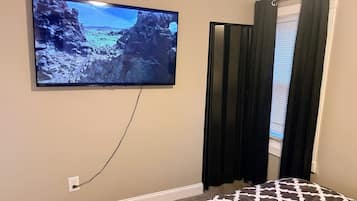 Una habitación con un televisor montado en la pared, una ventana con cortinas oscuras y una alfombra estampada.