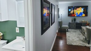 Une salle de bain moderne avec un lavabo blanc, un mur d’accent vert, des œuvres d’art encadrées et un téléviseur à écran plat.