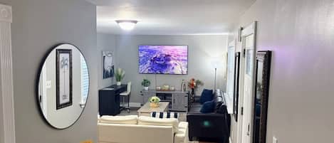 Un salon moderne avec un canapé, une table basse et un téléviseur. Un miroir est accroché au mur et une porte donne accès à une autre pièce.
