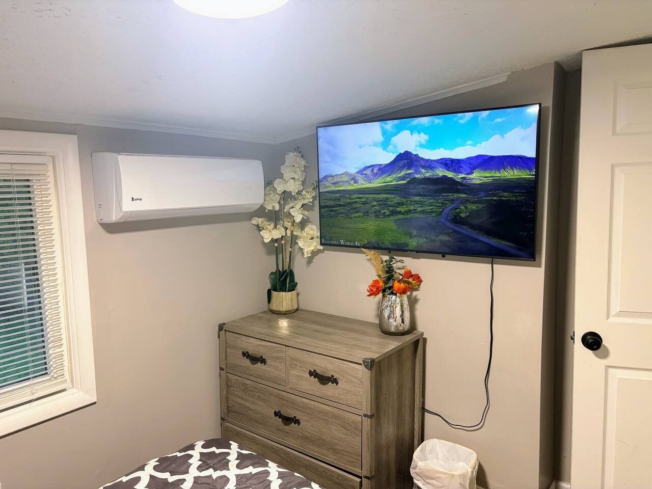A bedroom with a bed, a dresser, a television mounted on the wall, and a window with blinds.