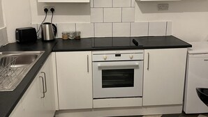 Una cuina compacta amb armaris blancs, taulell negre, forn de sobre, rentapelats i nevera petita.
