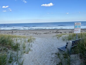 A beach path leading to the ocean with a signboard and a bench.