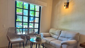 A cozy living area with a sofa, two wicker chairs, a small round table, and a large window with a view of greenery outside. - Natura Hotel y Villas  (Zihuatanejo)