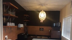 A compact kitchen with a black countertop, two stools, a hanging light fixture, and shelves with kitchenware. - Lovely Apartment in East London, South Africa (East London)