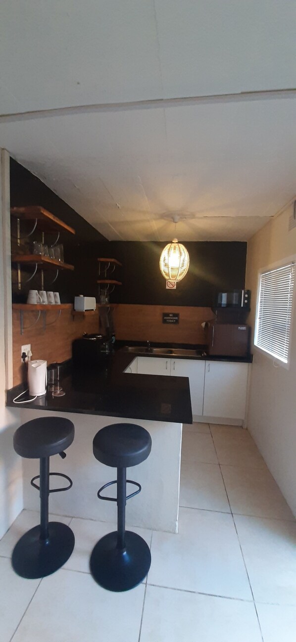 A compact kitchen with a black countertop, two stools, a hanging light fixture, and shelves with kitchenware.