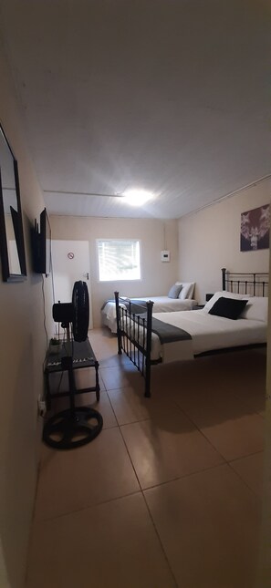 Quarto com cama, escrivaninha com ventoinha, janela com persianas e televisão fixada na parede.