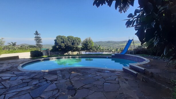 A swimming pool with a stone deck, surrounded by greenery and a view of hills. - Lovely Apartment in East London, South Africa (East London)