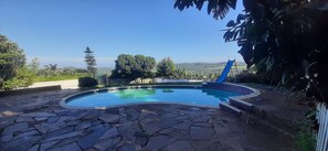 A swimming pool with a stone deck, surrounded by greenery and a view of hills.