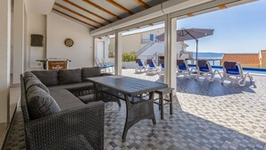 Un espacio de estar al aire libre cubierto con mobiliario de mimbre, una mesa y sillas, con vista a una piscina y un edificio con techo de tejas.