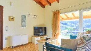Una habitación acogedora con techo de madera, un televisor sobre un mueble blanco, una ventana con cortinas y vistas al exterior con vegetación.