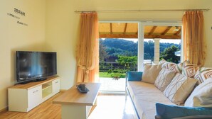 Sala de estar con televisor sobre un mueble de madera, sofá con cojines estampados, una mesita y vista a un jardín a través de una puerta corredera.