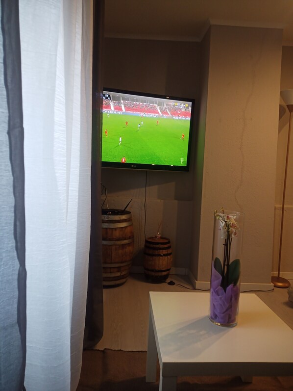 A room with a television mounted on the wall displaying a sports event, a wooden barrel, a vase with flowers on a table, and a curtain partially drawn.